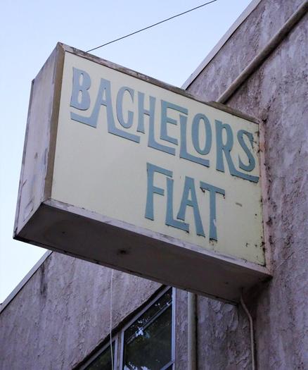 Backlit sign attached to a pinkish stucco building that says "Bachelors Flat" in a very swanky, 1960s style teal font.