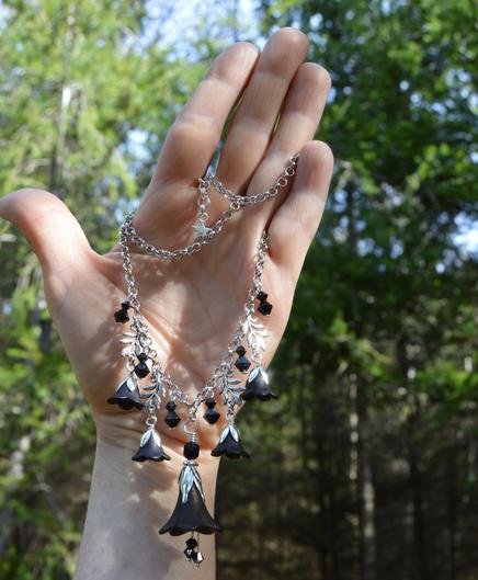 A black flower crystal necklace displayed hanging from an upright open hand. The necklace is V shaped, with a large black flower, and smaller ones hanging either side, along with black diamond shaped crystals and silver leaves. The background is a dark forest dappled with beright sunshine.