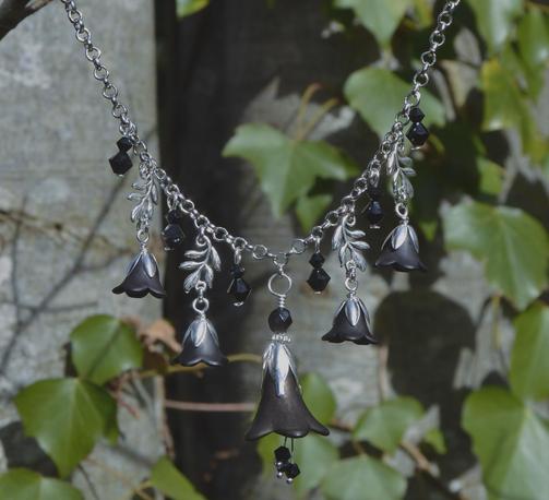 Close up of a black and silver necklace of flowers. A loarge central flower, tulip shaped, with 3 small crystals hanging underneath. Either side, smaller flowers, leaves and crystals dangle from a silver chain of round o shaped lings. Background is grey tree bark covered in ivy.