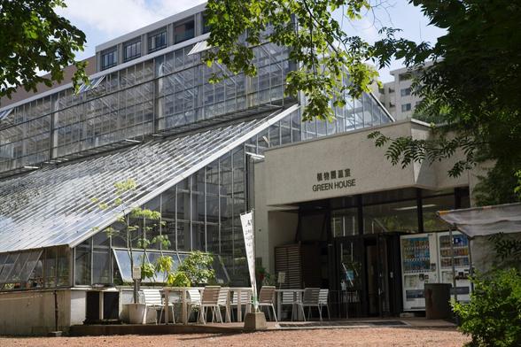 Entrance to a green house. Large slanted glass building with some patio furniture in front and 2 vending machines.