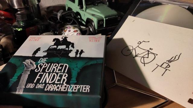 CD-Box des Hörbuches: Die Spurenfinder und das Drachenzepter. Daneben eine Kritzelzeichnung mit Fahrrad und einem Tisch mit Kaffeepott.