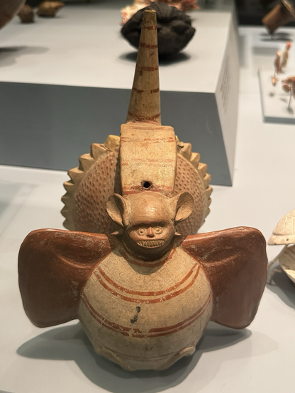 photo of the ceramic bottle on display at museum, front view showing bat figure with shell figure partially visible behind and spout in back bat with round body, spread open wings, and face that resembles the 😬 emoji!  light brown base color with darker red-brown painted details