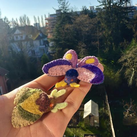 Two little felt moths held in my hand, overlooking some gardens. One moth has open wings. It is purple with yellow highlights. The smaller one has closed wings and is toned town yellow, brown and green.