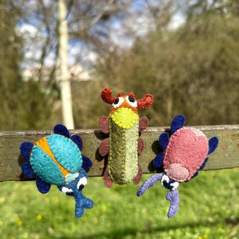 Three felt beetle magnets stuck to a metal fence in a park. One is blue and orange, another is long, thin and green and orange, and the third one is pink and blue.