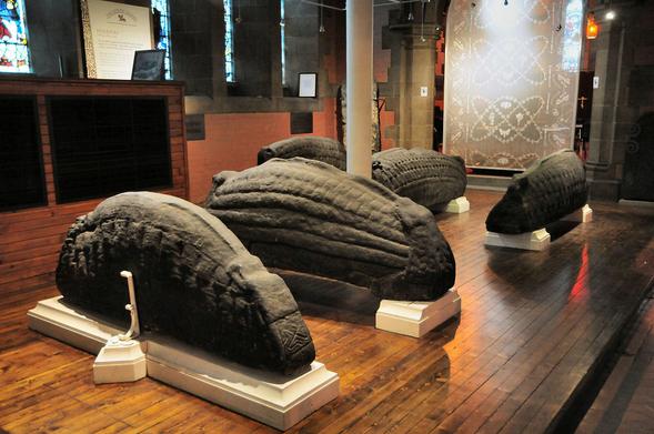 Viking hogback gravestones. The image shows part of the interior of a church. Five stone hogback gravestones are positioned on plinths on the floor. The two nearest are seen almost side-on while the further three are almost end-on. There’s a decorated screen in the background.