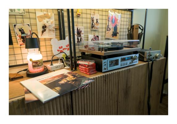 Photo showing the audio corner in my home office, with an Audio Technica record player (model AT-LPW40WN) standing atop a Pioneer 80s HiFi cassette player (model CT-300), with Stax Headphones (or "ear speakers," as the company likes to call them) with accompanying amplifier (models SR-404 and SRM-006t, respectively). To the side of the cassette player is a stack of five blank cassette tape cases, four of which are still shrink wrapped. The fifth is currently in the player, which has a small red light on indicating it is currently recording. Further to the left of the blank cassettes the "Midnights" LP album by Taylor Swift is laying, right in front of a candle light that is turned on. On the wall behind all this a metal wireframe on which a handful of photos are attached. Most every photo is of Yamgom, our black chow-chow mix dog in various poses and wearing various accessories. The overall mood of the photo gives me a sort of 90s nostalgic vibe, in large part due to the prevalence of physical objects that in the modern day more often than have become fully digital (photos, music).
