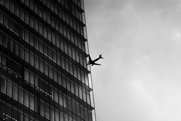 The image depicts a black and white scene where the silhouette of an airplane appears to be flying close to, or possibly through, a tall glass building. The stark contrast and composition create a dramatic effect.