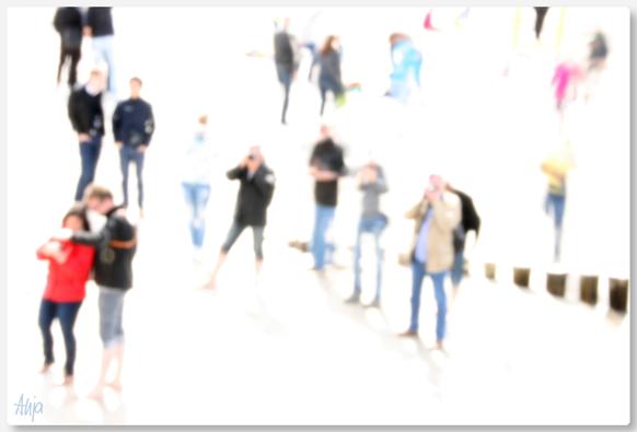 Eine Gruppe von Menschen am Strand, sie fotografieren etwas am linken Bildrand oder sie schauen auf Kameramonitore oder sie sprechen miteinander. Alle Menschen sind unscharf fotografiert und sind nur noch schemenhaft zu erkennen. Viele tragen schwarz-graue Kleidung, nur ein paar Farbkleckse sind im Bild zu erkennen. Weder Strand noch Wasser sind zu erkennen, nur eine Reihe von Holzbuhnen zeigt, dass die Menschen am Strand stehen.

A group of people on the beach, they are photographing something on the left edge of the picture or looking at camera monitors or talking to each other. All the people are blurred and can only be vaguely recognised. Many are wearing black and grey clothing, only a few splashes of colour can be seen in the picture. Neither the beach nor the water can be recognised, only a row of wooden groynes shows that the people are standing on the beach.