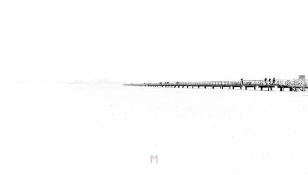 A high key black and white photo of the beach of Sankt Peter-Ording. The photo is mostly white. In the horizontal middle is a wooden walk way build on the beach. the way ends about in the vertical middle of the picture. There are some people on the walk way.