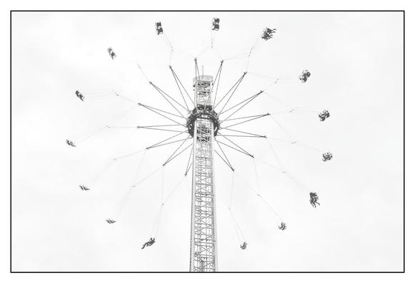 The picture shows a “high-key” black and white image of a “Star Flyer” carousel in motion, taken from below. In the center is a tall, slender metal tower that tapers towards the top. At the top of the tower is a rotating ring, from which numerous rows of seats swing outwards on long ropes. The seats are arranged in pairs and the passengers are suspended in the air as the carousel rotates, creating a dynamic, star-like effect. The sky in the background is bright and uniform, which draws the focus to the structure of the carousel. The lower part of the tower is not fully visible as the image is oriented upwards.