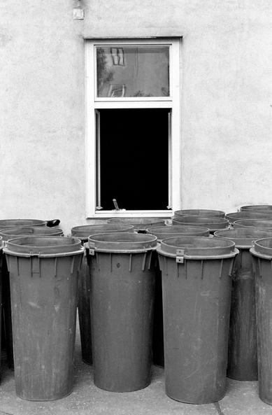 The black and white image shows a group of tall plastic trash bins lined up in front of a plain wall with a single open window. Reflections of buildings are visible in the top windowpane, while the bottom half is open, revealing darkness inside.