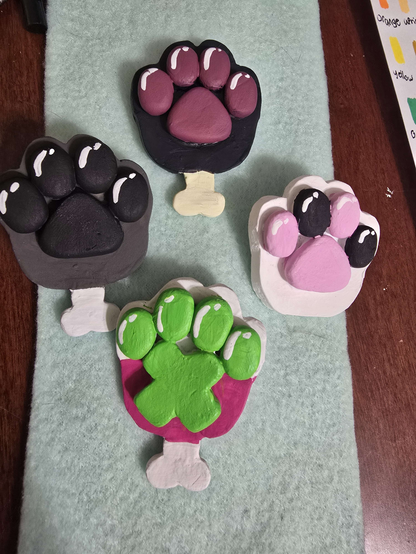 four polymer clay paw shaped pins, three have a little bone sticking out the bottom at the wrist, the one that doesnt is white with pink and black beans, one is a pink paw with white toes green beans and an X shaped pawpad, another is a dark brownish grey paw with a white bone and black pads, the last is a black paw with dark fleshy pink beans and a yellowish bone