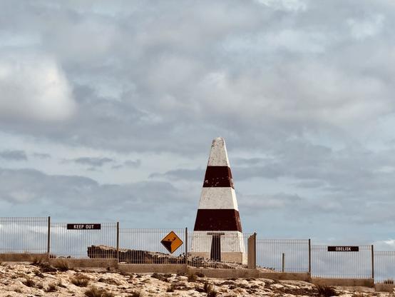 An alternating white and red obelisk sits beyond a fence with signs saying “KEEP OUT”, “OBELISK”, and a yellow sign warning of a falling danger. A cloudy sky is beyond. How many victims has it lured, we wonder?
