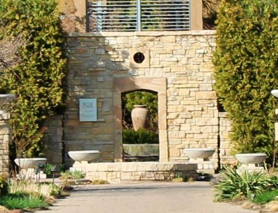 Paver walkway leading to short stone wall of an empty water feature in front of a stone structure with a doorway and a small circular open air window above the doorway. Four empty cement garden planters are in front of the structure and a metal railed observation deck is above. Evergreen trees are growing next to the structure. Through the doorway a large urn is standing in bright sunlight behind some green plants set behind a weathered wooden bench with a rail backrest with evergreens behind the urn.