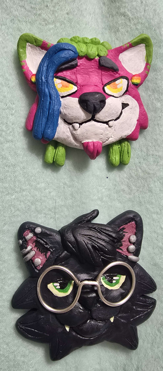 two polymer clay headshot pins, one of a pink hyena with green and blue locs, white muzzle and yellow eyes, the other a black cat with green eyes and glasses