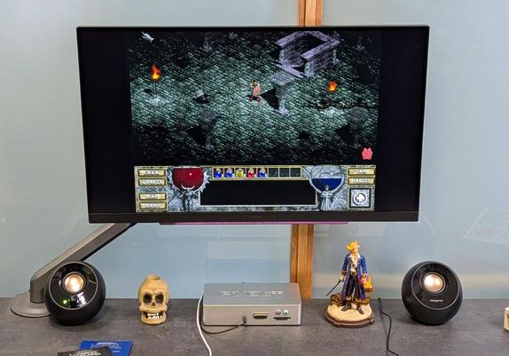 A computer monitor displays a retro video game with a medieval fantasy setting; speakers, a skull ornament, and a figurine sit on the desk below.