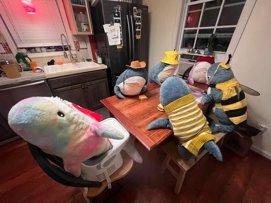 This image shows a whimsical scene of plush sharks (BLÅHAJ) set up at a kitchen table as if having a meal together. In the foreground is a large light blue and pink BLÅHAJ sitting on what appears to be a small stool. Around a wooden table are five more BLÅHAJ plushies dressed in various outfits - they're wearing blue jeans, and some have yellow hats or striped shirts. One is even dressed like a bee with black and yellow stripes. The sharks are positioned as if they're enjoying dinner together in a cozy kitchen setting with a refrigerator and sink visible in the background. The wooden floor and homey kitchen create a warm, domestic atmosphere for this charming stuffed shark gathering.​​​​​​​​​​​​​​​​