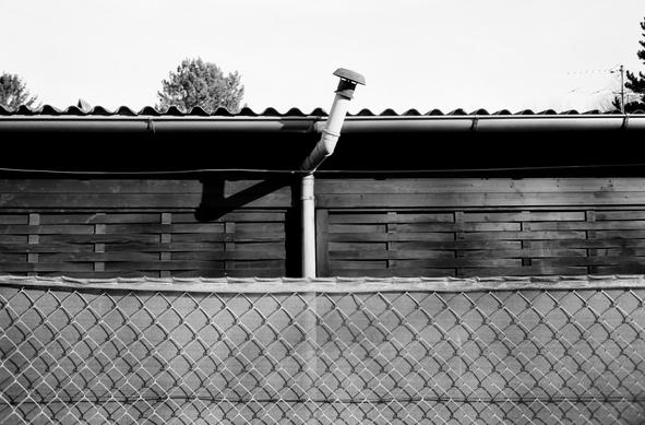 The black and white image shows a wooden building with a corrugated roof, a slightly tilted metal chimney, and a chain-link fence in the foreground, with a few trees visible in the background.