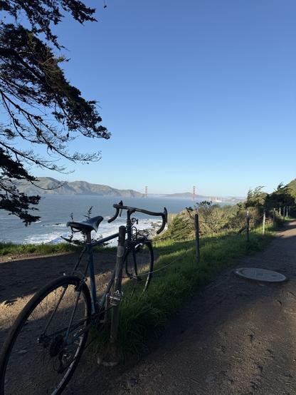 A bicycle is leaned against a fence along a dirt path with lush green grass. In the background, the Golden Gate Bridge spans across the water, under a clear blue sky. The coastline and ocean waves are visible on the left.