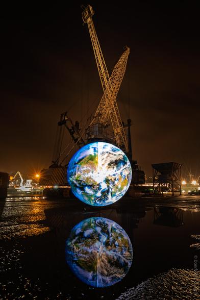 A striking night-time photograph taken in Cork, Ireland, featuring a large illuminated globe installation set in front of an industrial crane, with the glowing sphere beautifully reflected in a puddle on the ground; city lights and industrial structures are visible in the background