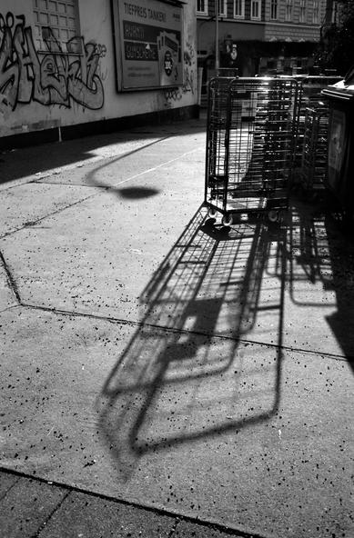 The black and white image shows a group of metal rolling carts casting long, distinct shadows on a sunlit concrete sidewalk, with graffiti-covered walls and a billboard in the background.