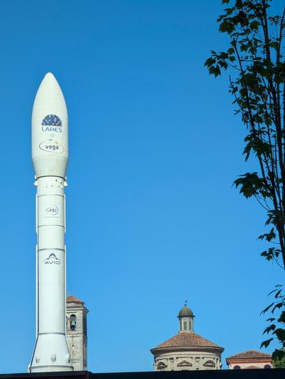 A daylight outside view with clear blue sky taking up most of the vertically oriented frame. A white rocket stands close to the left edge of the frame. The upper branches of a tree are partially seen along all the right edge. The upper parts of the main building and the belltower of a 16th century basilica are partly seen at the bottom of the frame.