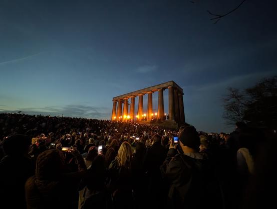 A crowd of people in front of the National Monument, an unfinished columned portico modeled after the Parthenon, where a line of robed figures hold torches at dusk.