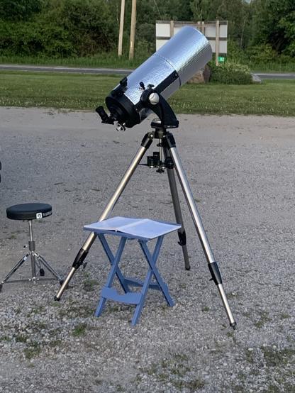 A picture of my telescope (a 1985 Meade 2080 LX3) sitting in a gravel parking area with my drum throne and a small table beside it. On the table are some paper star charts (paper like a pirate! Arrrr!).