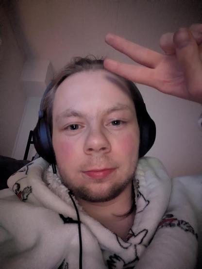 The image captures an adult person indoors. In the foreground, the person is wearing headphones and a patterned hoodie. A hanger is vaguely visible behind the person.