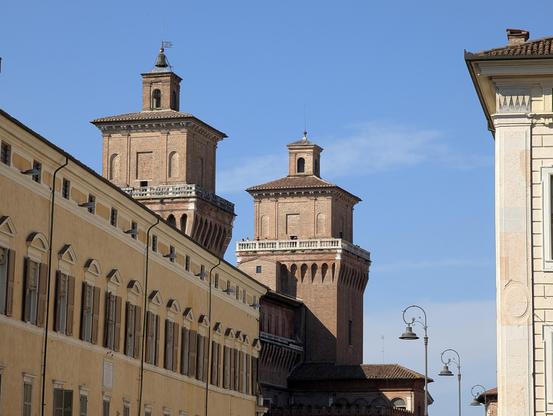 View of two medieval brick towers of the Castello Estense in Ferrara, Italy, rising above surrounding buildings under a clear blue sky. The warm tones of the architecture contrast with the crisp light, and vintage-style street lamps line the street below.