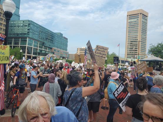A crowd of protestors on a plaza in downtown Baltimore. Most signs aren't fully legible but one says "Liberty and justice FOR ALL"
