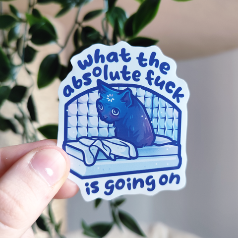 hand holding up a sticker of a cat waking up in psych ward titled "what the absolute f*ck is going on"