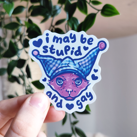 hand holding up a sticker of a cat wearing a silly hat titled "i may be stupid and gay"