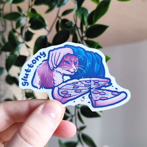 hand holding up a sticker of a cat being pulled away from eating a pizza titled "gluttony"
