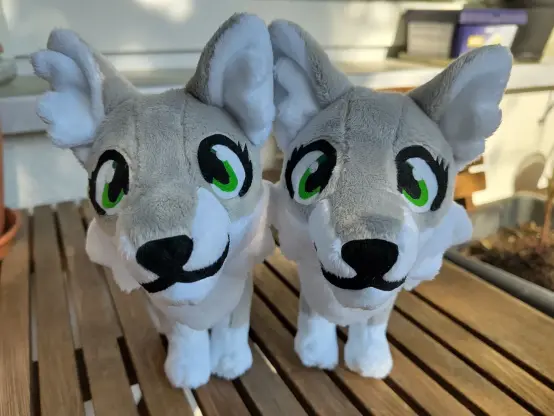 Two small grey wolf plushies with large green eyes sitting on a wooden table. Their heads look oversized from this perspective and they seem to by saying "buy me!".