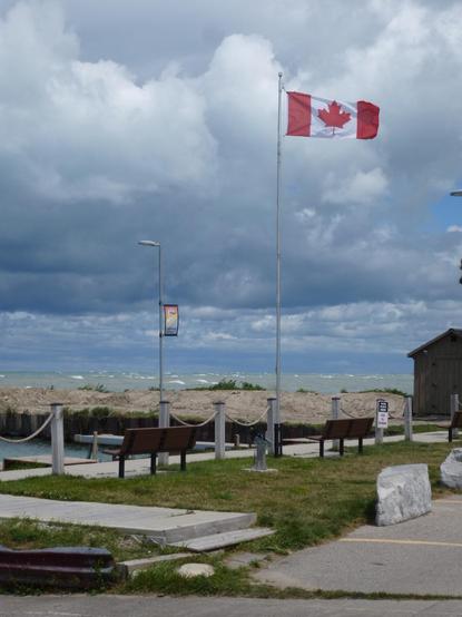 A Canadian flag blows in the wind, a large waved covered lake can be seen in the distance. The sky is covered in large dark clouds and benches and rocks can be seen in the foreground.