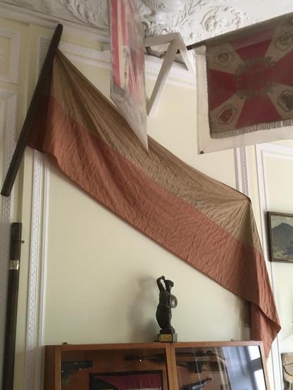 long, faded Polish flag hangs down at a slight diagonal from left to right against a light-colored wall

at top right, a square military flag hangs from a horizontal rod at ceiling level

on top of a display case below, a small sculpture of the famous Warsaw Mermaid  https://www.atlasobscura.com/places/mermaid-of-warsaw

more images from the Museum here
https://pism.org.uk/sikorski-museum/