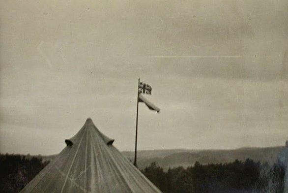 black-and-white photo, landscape orientation

left foreground, the pointed top of a round tent
at right, a crude flagpole from a young tree: British flag above the Polish
background: forested hills