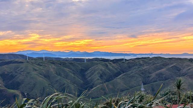 Looking west from Makara Peak over green hills, ocean and land in the distance with the sun low in the sky.