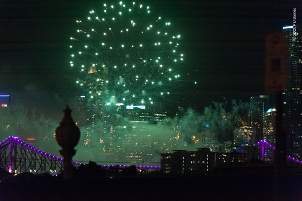 A green ball of fireworks explodes over the drifting smoke of previous pyrotechnics. Below and behind are the outlines of city buildings and the purple lights of the Storey Bridge.