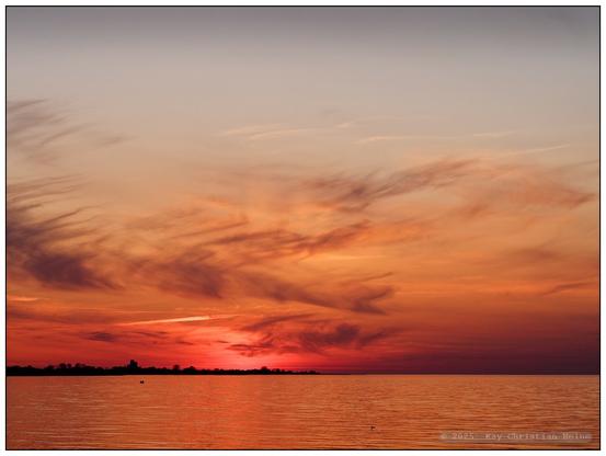 Sonnenuntergang über der Ostseeküste bei Hohenfelde im nördlichen Kreis Plön mit flammend orange-rotem Himmel und ruhigem Wasser im Vordergrund, am Horizont von links bis zur Bildmitte ein schmaler, silhouettenhaft schwarzer Streifen Landes.