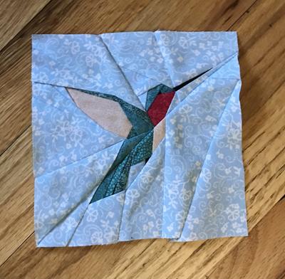 Hummingbird quilt block. The bird is in green fabrics with a ruby throat. The background is a light blue and white floral print.
