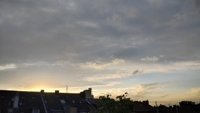 Foto im Querformat 16:9. Blick über die Hausdächer von Mietshäusern. Der Himmel darüber ist von der untergehenden Sonne leicht gelb. Die Sonne selbst lugt noch ein kleines Stück hinter einem Dach links hervor. Weiter oben hellgraue Wolken.