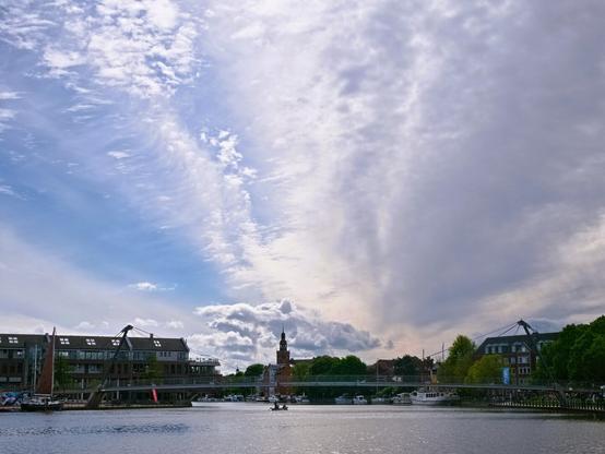 In the foreground, the water of a harbour. Buildings, trees and boats to the left and right. A curved bridge spans the water. A tower can be seen on the horizon. And above it a lot of sky with white cloud mountains, a denser cloud cover and blue patches.
