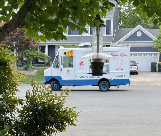 Photograph of an ice cream truck painted white and blue, stopped in a suburban cul de sac with a two story colonial house behind it.