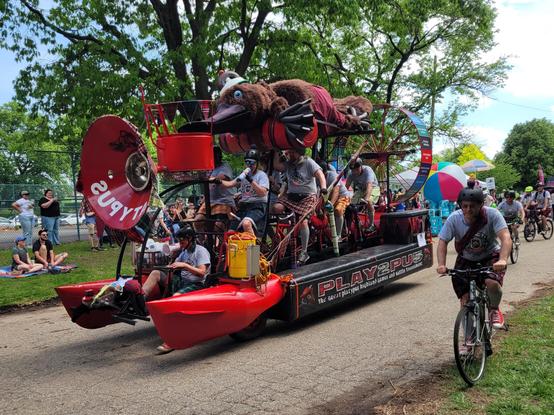 Amphibious human powered vehicle titled play2pus, made of a metal frame connecting bicycles over a red pontoon base, topped by a giant stuffed platypus. The front of the vehicle is decorated as an air horn with "platypus" written backwards like an ambulance. A price is right wheel sits on the back of the vehicle, listing highland games to try. The riders are costumed in kilts and Scottish hats, as is the platypus.