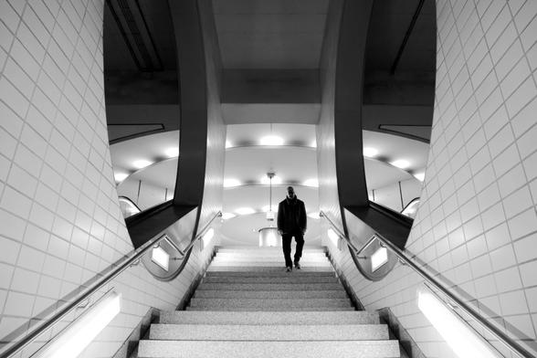 black and white. portrait format. the staircase of a subway station in frankfurt. architecturally very striking. the view leads from the bottom to the top. the walls are brightly tiled, illuminated and have a large circular recess on the left and right. the ceiling of the upper level, which is also brightly illuminated, can be seen in the background. The lighting is also arranged in a circle and harmonizes with the round recesses in the walls. a man walking down the stairs appears as a dark silhouette.