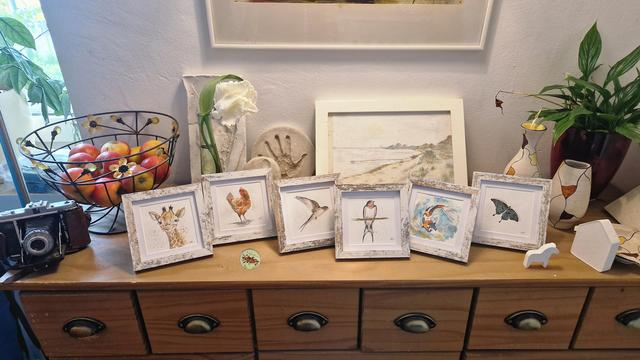 Auf einer Holzkommode stehen insgesamt 9 handgemalte Bilder, 6 kleinere quadratische Tier Illustrationen jeweils mit dem Motiv , Koi Fisch, zweimal Schwalbe ,  grüner schwalbenschwanz-schmetterling, Giraffe,  Huhn und dahinter  eine unverkäufliche landschaftszeichnung der SchleiGegend.