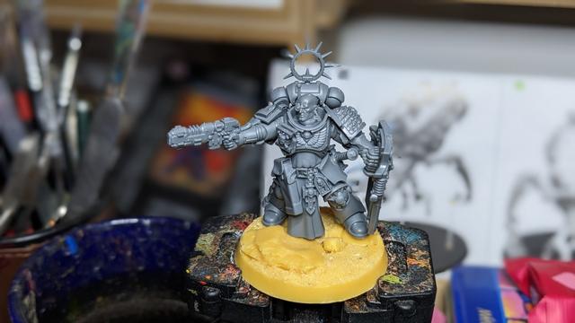 An unpainted grey Space Marine Captain with Deathwatch shoulder pad and shield standing on a yellow base.