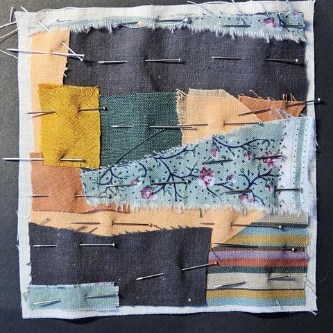 Ein paar Stoffschnipsel, die mit Stecknadeln für eine Stoffcollage auf ein Stück Untergrundstoff aufgepinnt sind.